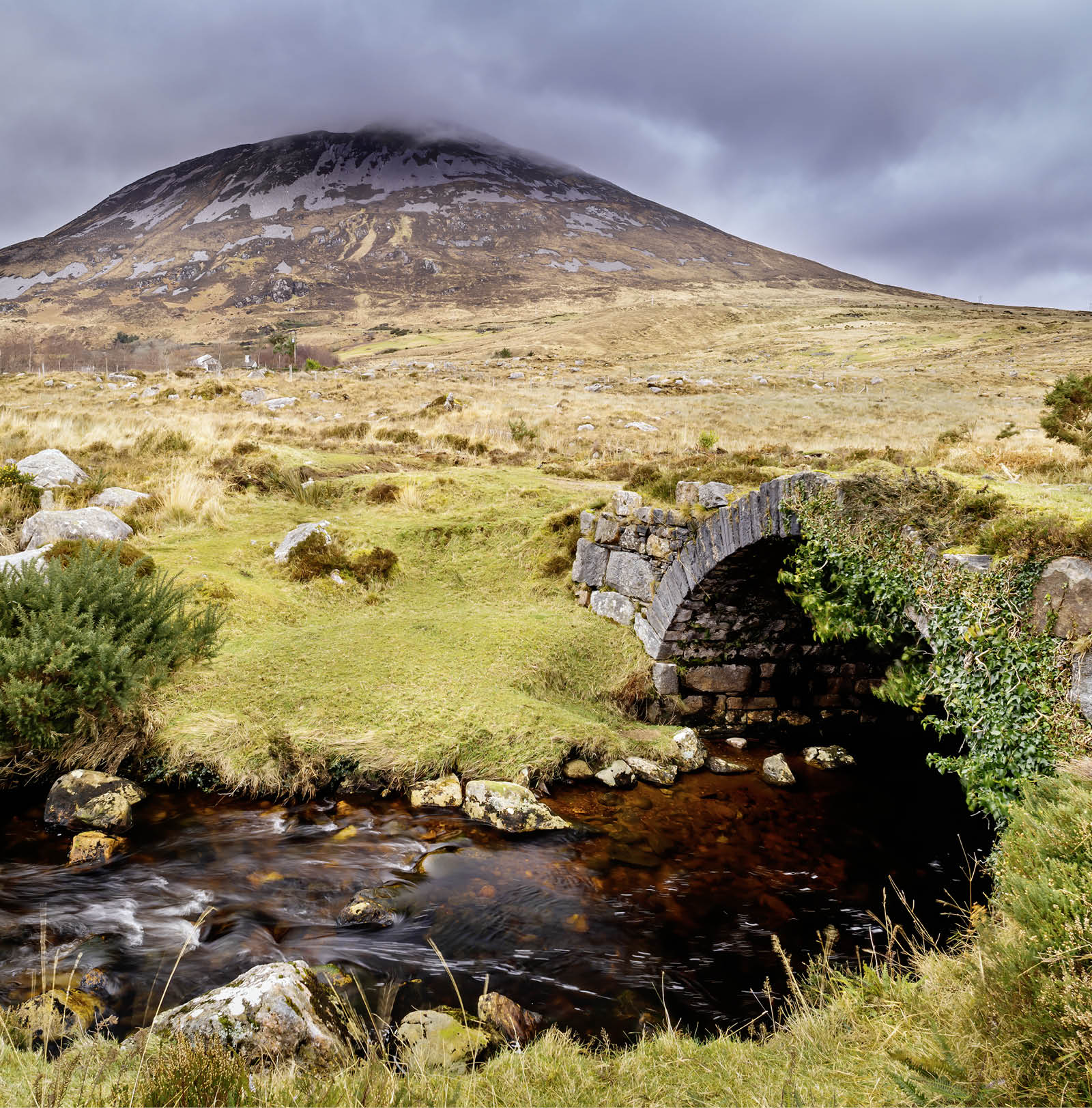 Old small stone bridge over river with view at Errigal mountain and stormy clouds, County Donegal, Ireland