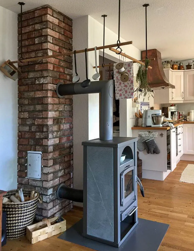 Ein Kaminofen aus Antik Classic Backsteinriemchen wirkt klassisch und fügt sich im Gesamtbild des Raums toll ein. Die weiße Küche und der Holzboden passen perfekt zur Kaminrückwand aus rot braunen Riemchen