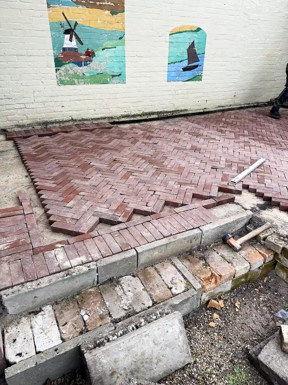 Roter Pflasterklinker der gerade im Fischgrätmuster verlegt wird. Dahinter eine gelbe Wand.