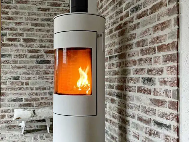Weißer angefeuerter freistehender Kamin. Er steht auf einem Klinkerboden und die Wände hinter dem Kamin sind auch verklinkert.