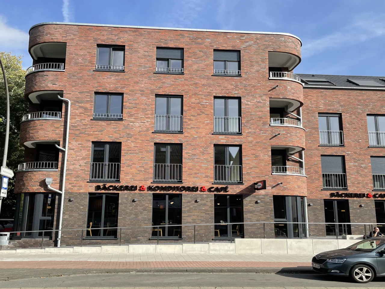 Viergeschossiger Flachdachbau mit roten glatten Klinkerriemchen ab dem ersten Geschoss. Das Untergeschoss ist mit einem braunen Klinker verkleidet. Viele Fenster.